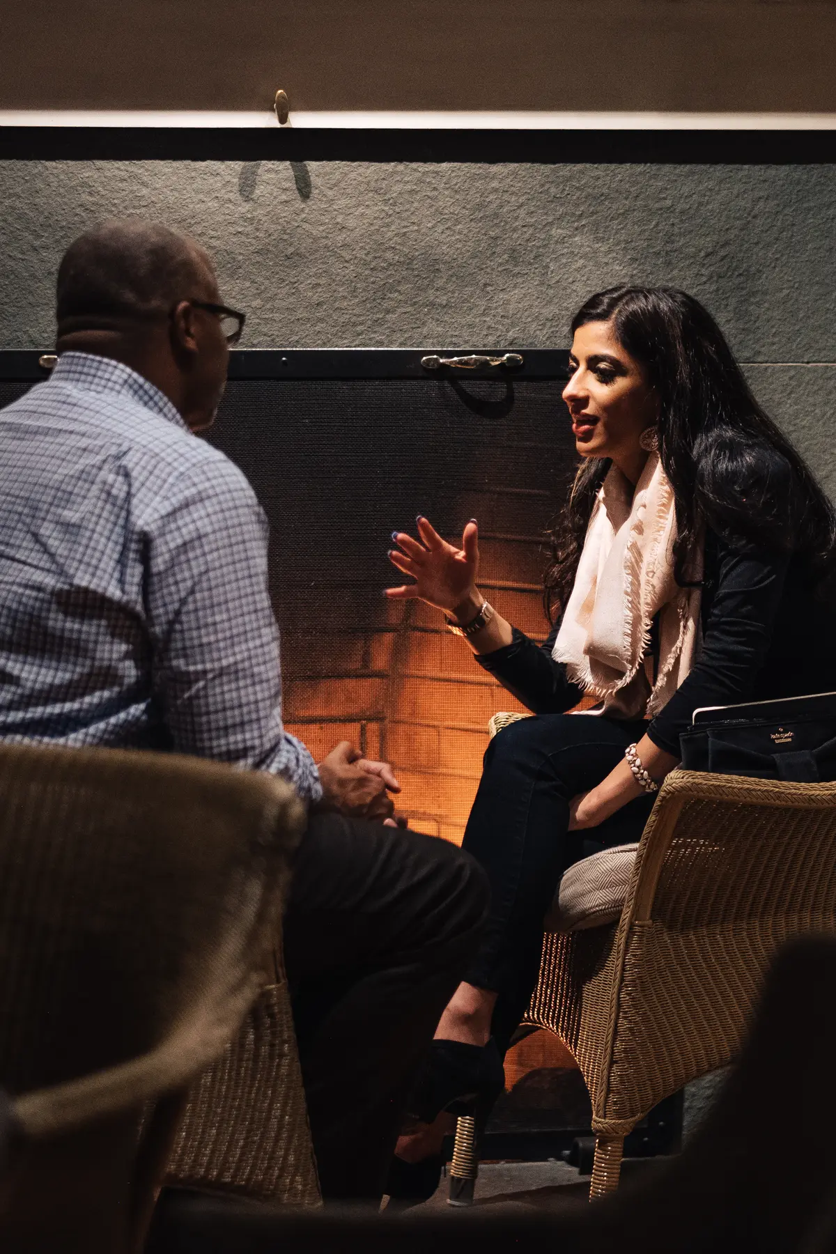 Two people engaged in a deep conversation while seated in wicker chairs near a warmly lit fireplace. The woman, dressed in black with a light pink scarf, gestures expressively as she speaks, while the man in a checkered shirt listens attentively. The cozy, dimly lit setting suggests an intimate or meaningful discussion in a relaxed atmosphere.