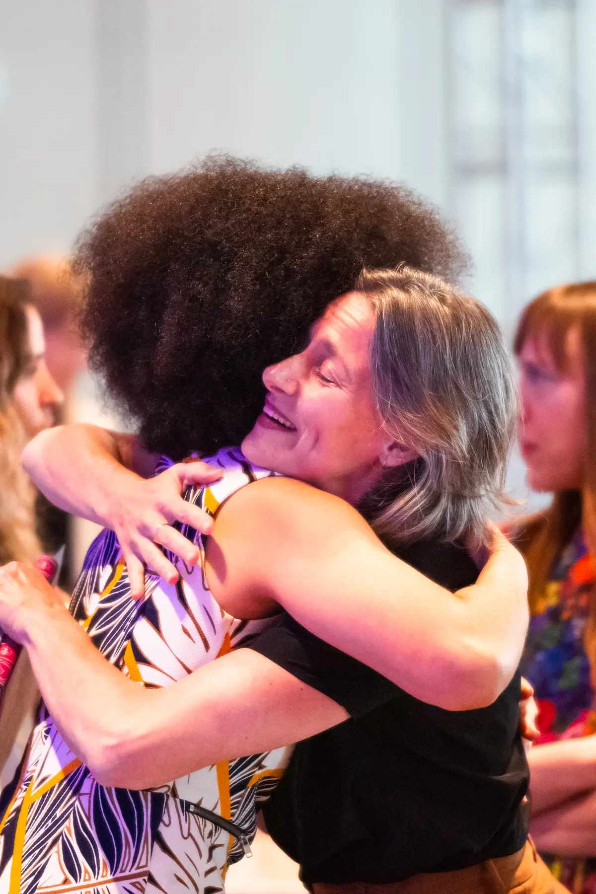 Two women embrace warmly at a networking event, expressing joy and connection. One woman, holding a microphone, wears a patterned sleeveless dress, while the other, in a black top, smiles with her eyes closed. In the background, two other women engage in conversation, adding to the lively and welcoming atmosphere of the gathering.