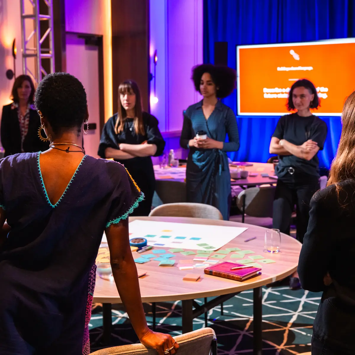 A group of people engaged in a collaborative workshop in a modern conference setting. A woman in a blue dress stands at the center, addressing participants who are gathered around a table covered with sticky notes and papers. Some attendees listen with arms crossed, while others engage in discussion. The background features a large screen with a bright red presentation slide and deep blue curtains, adding to the professional yet creative atmosphere.