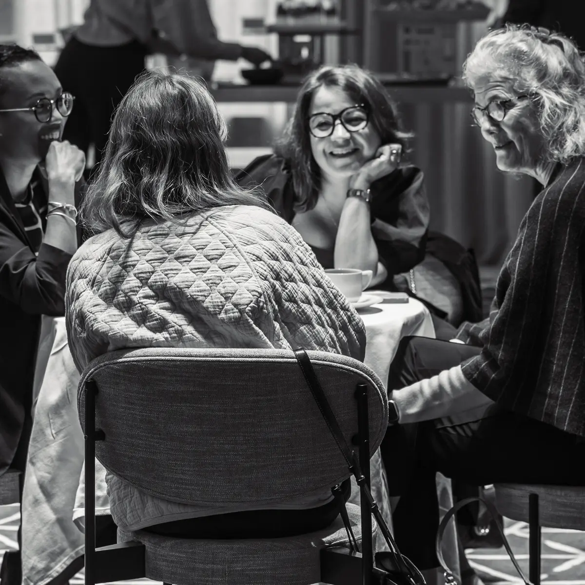 A black-and-white photograph of four women engaged in a lively conversation at a round table during a networking event. They are smiling and leaning in as they talk, creating a warm and engaging atmosphere. The background features a buffet table with refreshments, suggesting a relaxed yet professional gathering.