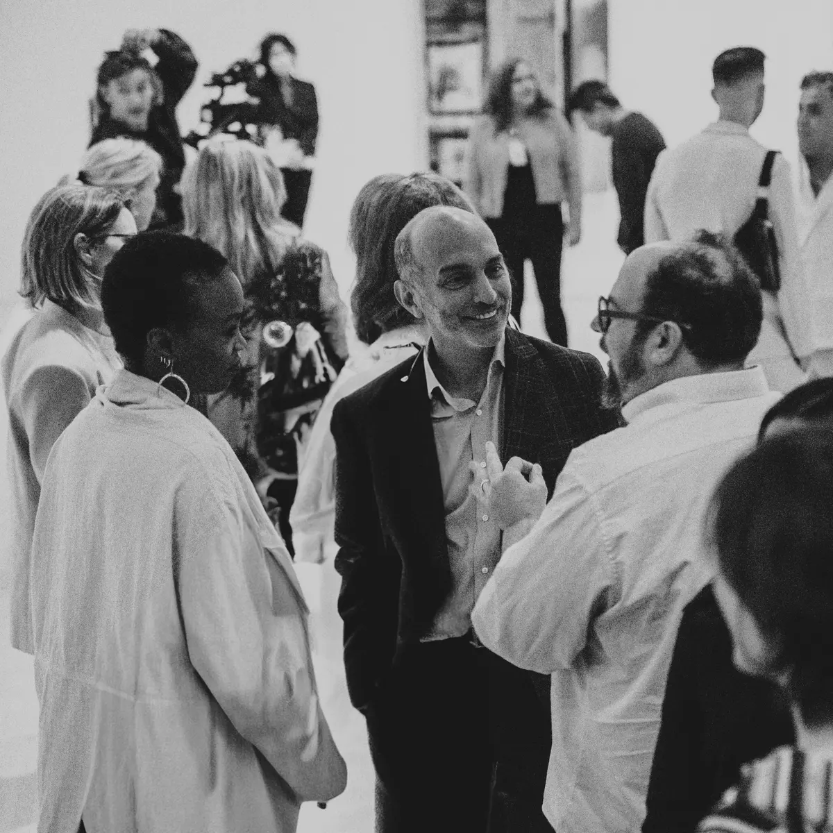 A black-and-white photograph capturing a lively networking event in an art gallery or modern space. A diverse group of people is engaged in conversation, with a man in a suit smiling as he speaks with another man wearing glasses and a white shirt. In the background, other attendees mingle, some observing artwork on the walls while a photographer captures the moment. The setting conveys a sophisticated yet relaxed social atmosphere.