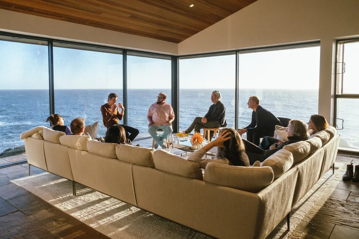 A group of people engaged in a thoughtful discussion in a modern living space with floor-to-ceiling windows overlooking the ocean. Seated on a large, neutral-toned sectional sofa and stools, they are illuminated by warm natural light. The serene coastal backdrop adds to the relaxed yet intellectual atmosphere of the gathering.
