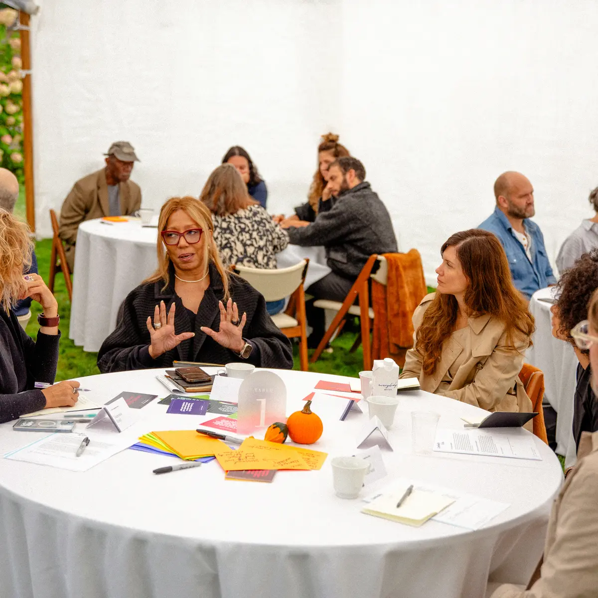 A diverse group of people engaged in a discussion at a round table under a white event tent, surrounded by greenery. A woman in glasses and a black sweater gestures as she speaks, while others listen attentively. The table is covered with notes, documents, colorful index cards, and a small decorative pumpkin. Additional participants are visible in the background, seated at other tables, fostering a collaborative and engaged atmosphere.