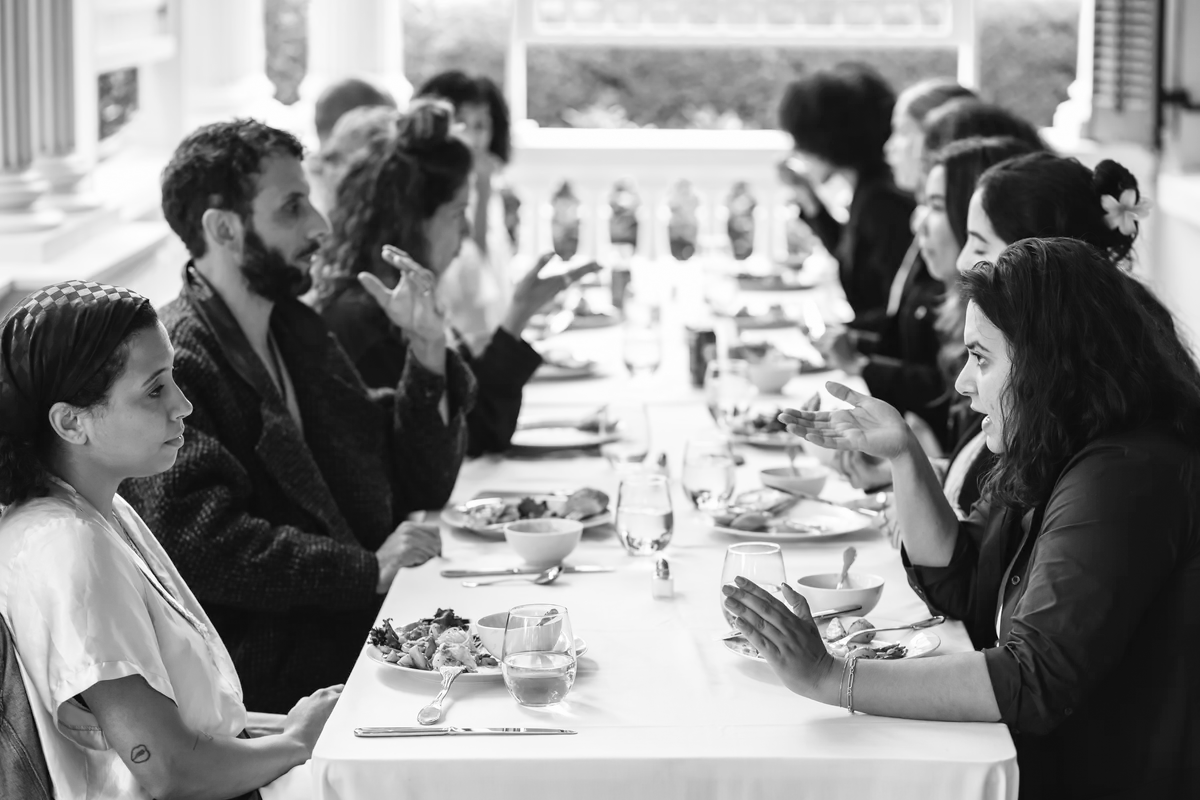 A group of people engaged in animated conversation over a meal at a long table covered with a white tablecloth. Two individuals in the foreground gesture expressively while discussing, while others listen and interact. The outdoor setting features a bright veranda with white railings and lush greenery in the background, creating a welcoming and convivial atmosphere.