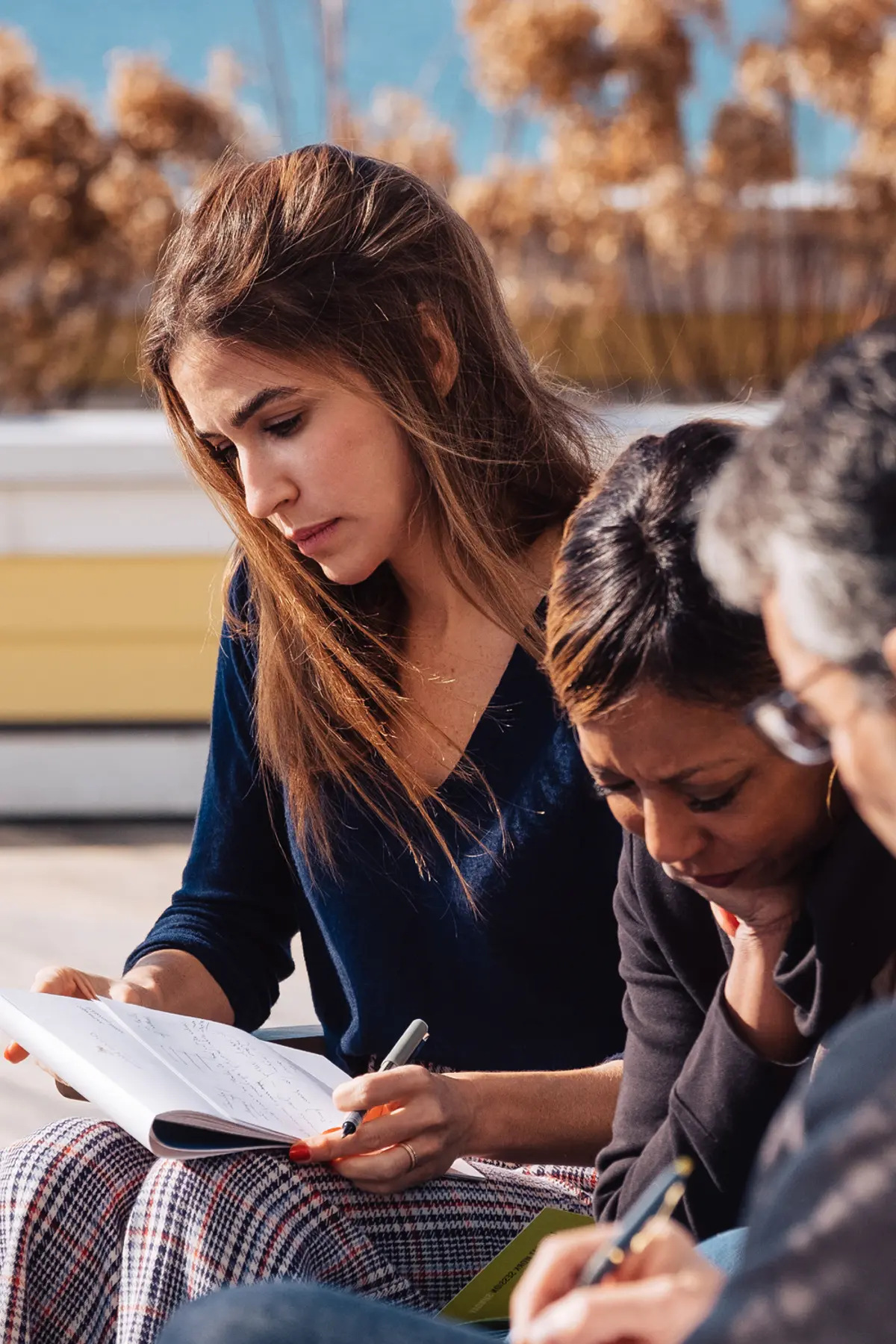 Three professionals engaged in an outdoor brainstorming session. A young woman in a navy blue sweater intently reviews notes in a notebook, while two colleagues, one in a black blazer and another in glasses, focus on their writing. The setting, featuring dried plants and a bright sky, suggests a thoughtful and collaborative atmosphere.