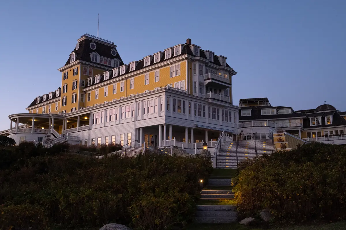 Grand historic hotel with yellow siding and white trim, illuminated at dusk with stairway lighting and a landscaped path leading to the entrance under a clear evening sky.