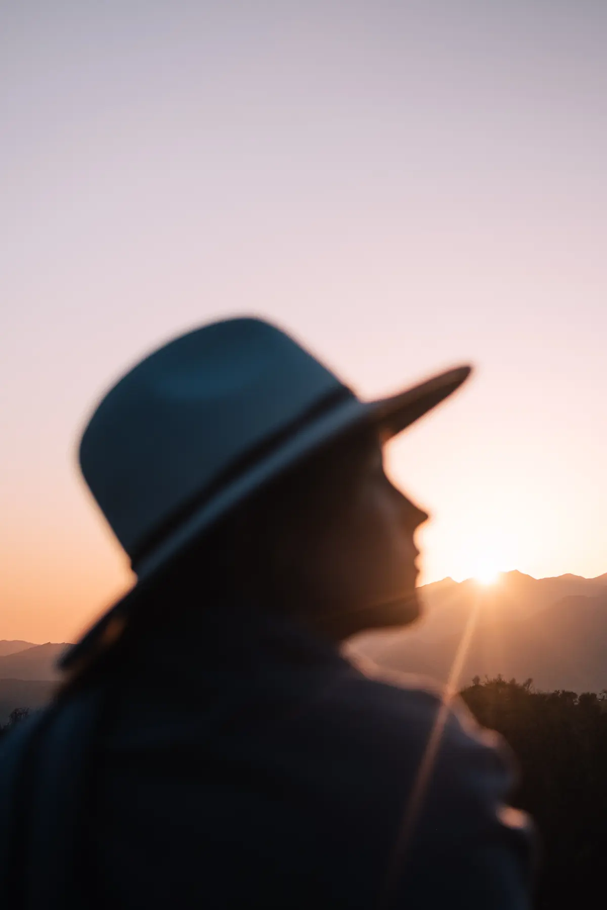 Silhouette of a person wearing a wide-brimmed hat watching the sun rise over misty mountain peaks, creating a tranquil and reflective moment in nature.