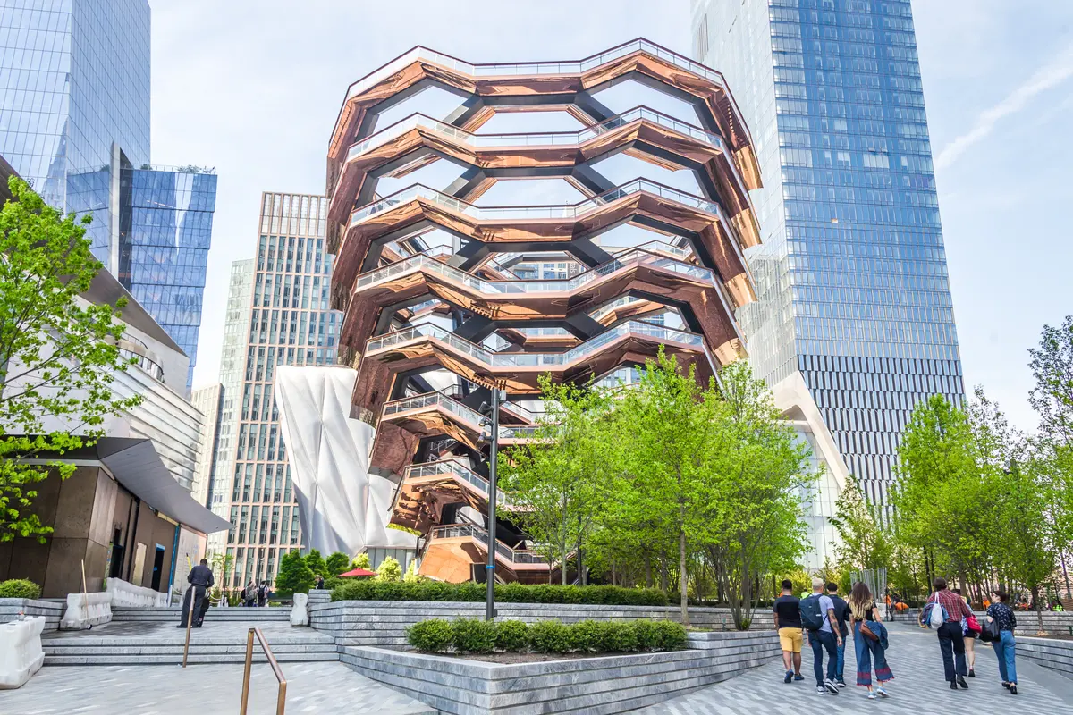 The Vessel at Hudson Yards in New York City, a striking honeycomb-like staircase structure surrounded by modern skyscrapers and lush green trees, with visitors walking along the plaza beneath a clear sky.
