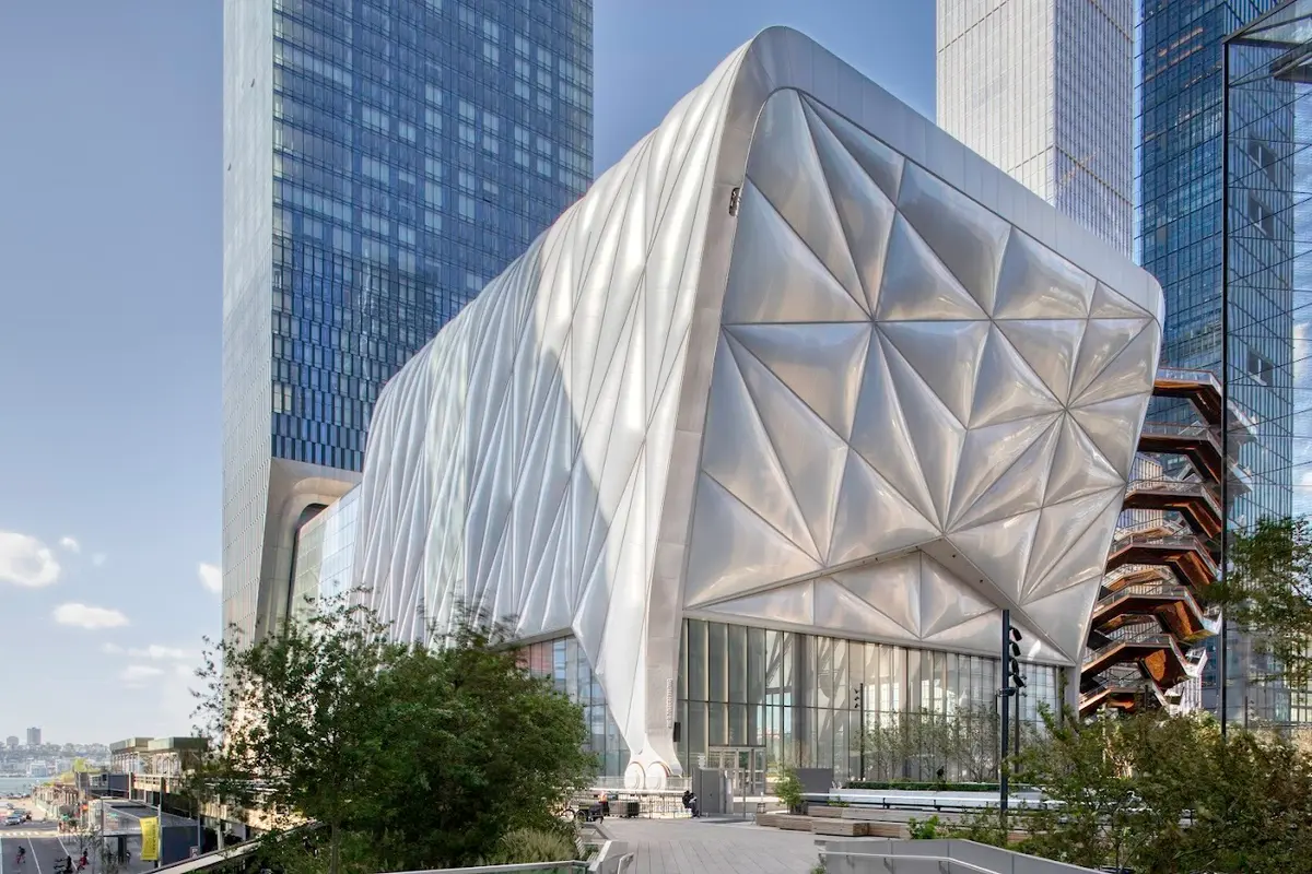 Futuristic view of The Shed arts center in New York City, featuring its distinctive expandable, metallic shell-like facade with geometric patterns, surrounded by tall glass skyscrapers and The Vessel sculpture; photo by Brett Byer.