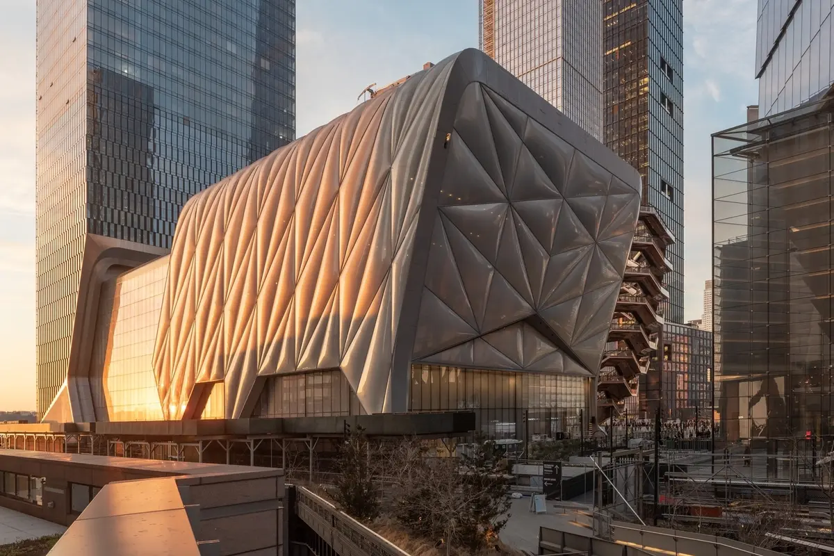 The Shed, a contemporary cultural center in New York City’s Hudson Yards, featuring its distinctive quilted, movable facade illuminated by the golden light of sunset, surrounded by modern glass skyscrapers.