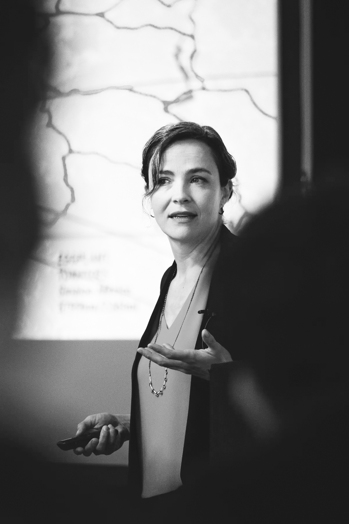 Black and white photo of a woman presenting in front of a projected map, speaking to an audience with a thoughtful expression, while holding a presentation remote.