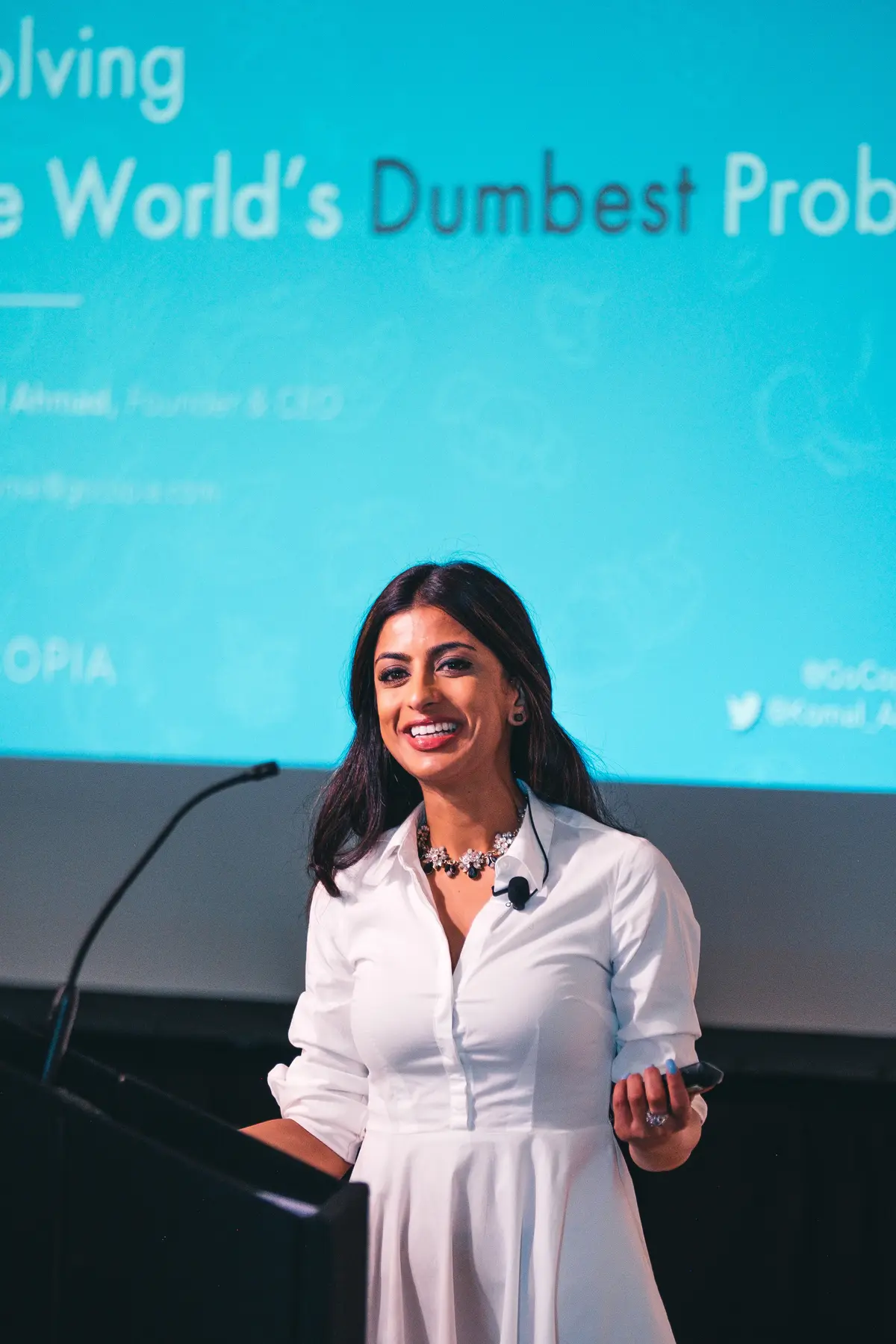 Komal Ahmad, Founder and CEO of Copia, smiles while speaking on stage in a white dress, standing at a podium in front of a bright blue presentation slide titled “Solving the World’s Dumbest Problem,” during a conference talk.