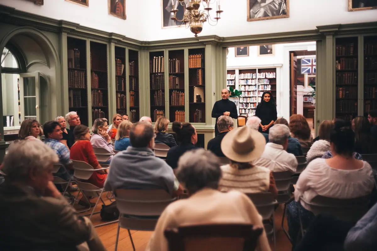 Two speakers address a seated audience in a historic library filled with antique books and classical paintings. The room is warmly lit, creating an intimate and thoughtful atmosphere as attendees listen attentively to the presentation or discussion.
