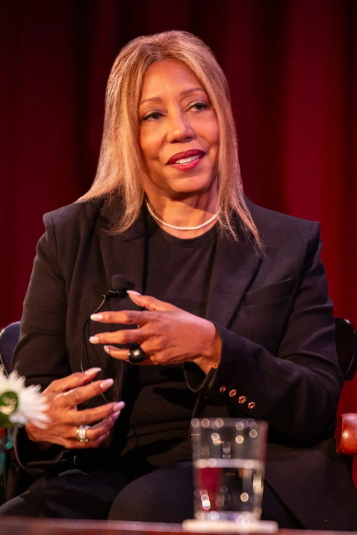 Denise Young speaks on stage during a civic imagination salon, seated in front of deep red curtains. She gestures thoughtfully while holding a microphone, wearing a black blazer and pearl necklace. A small table with a vase of flowers and glasses of water is visible in the foreground, adding to the intimate, conversational atmosphere.