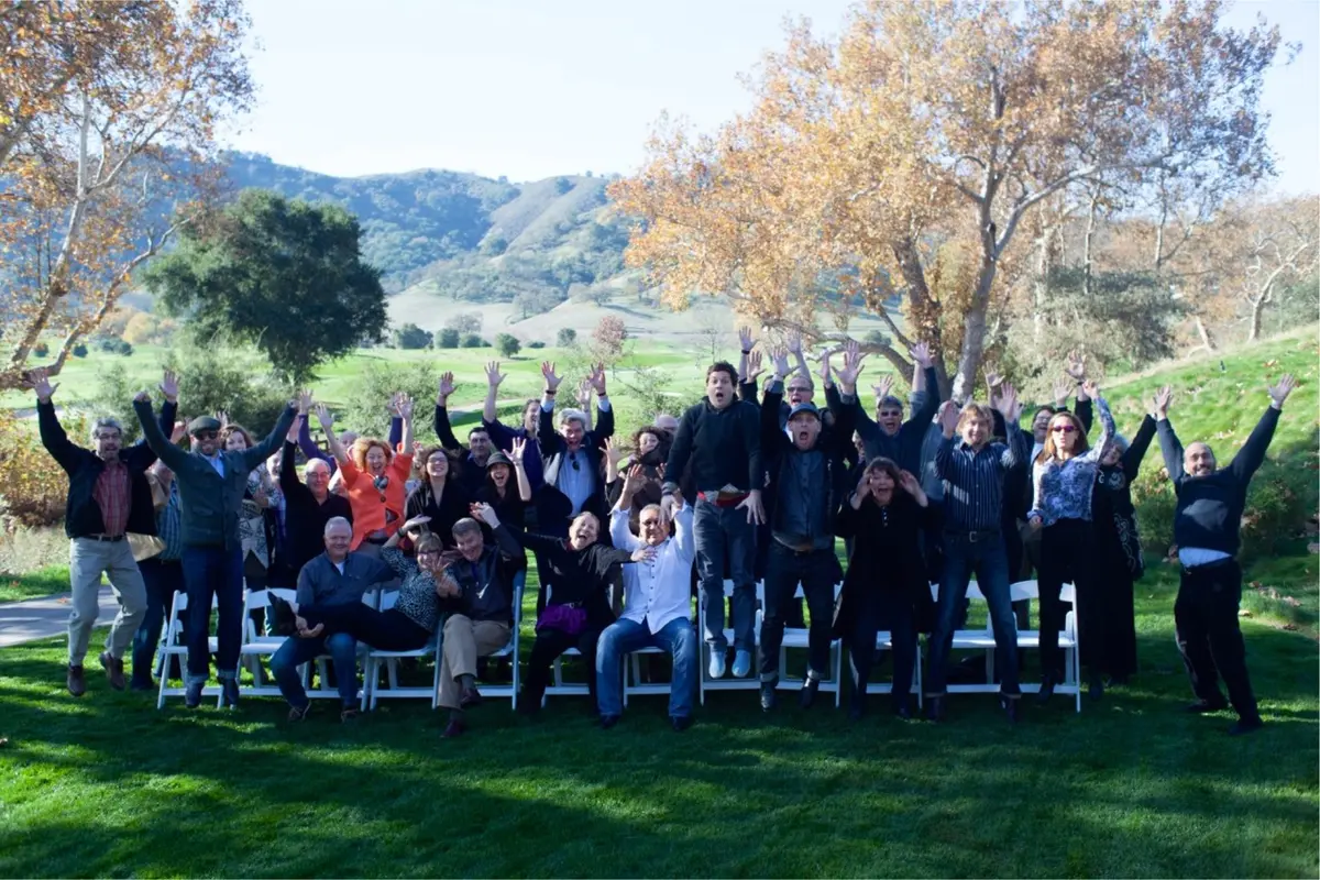 A joyful group of adults poses outdoors with arms raised in celebration, some mid-jump, against a scenic backdrop of rolling hills, trees, and blue sky on a sunny day.