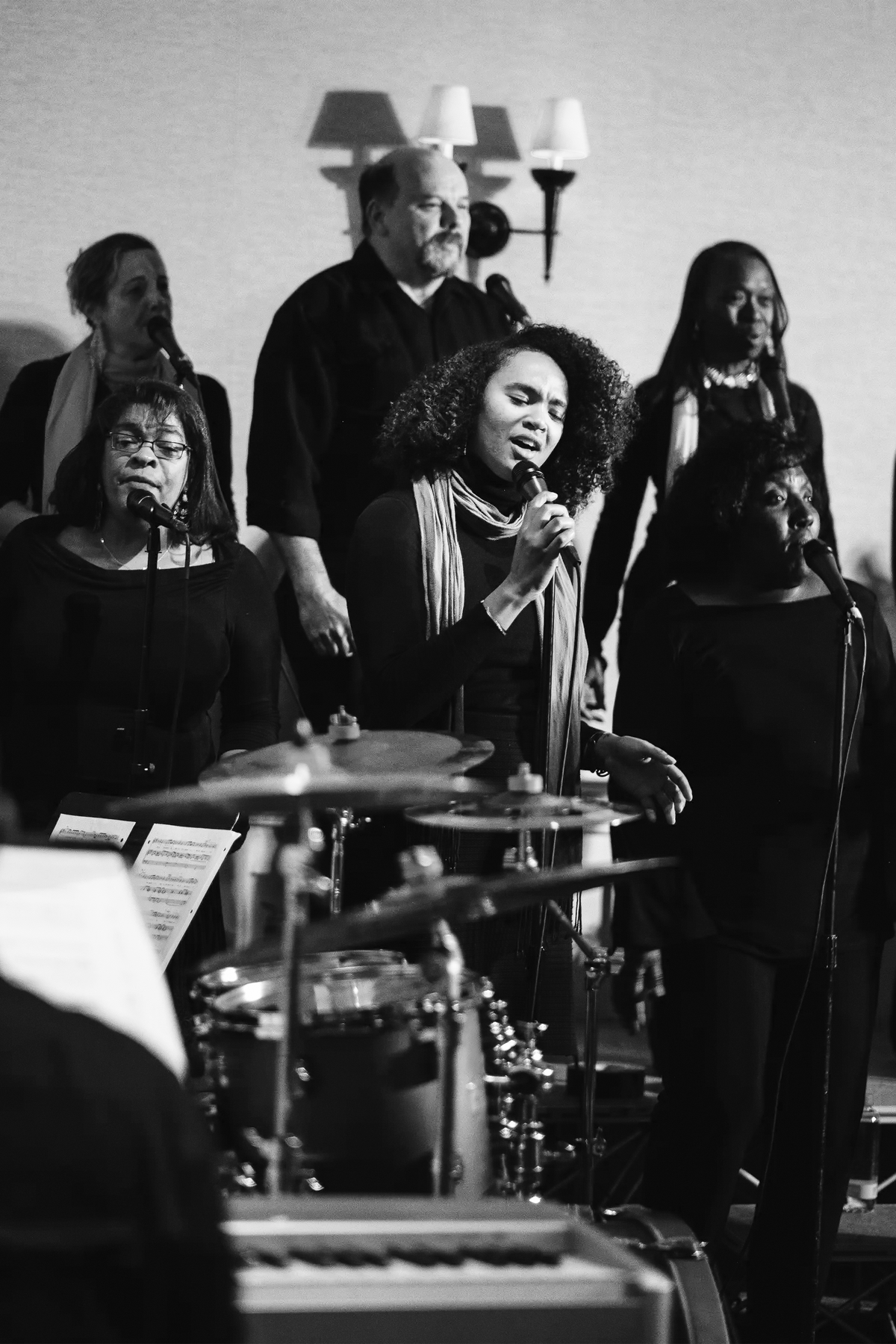 A diverse choir passionately performs on stage, with a woman in the foreground singing into a microphone beside a drum set, surrounded by fellow vocalists in black attire and colorful scarves, under warm stage lighting.