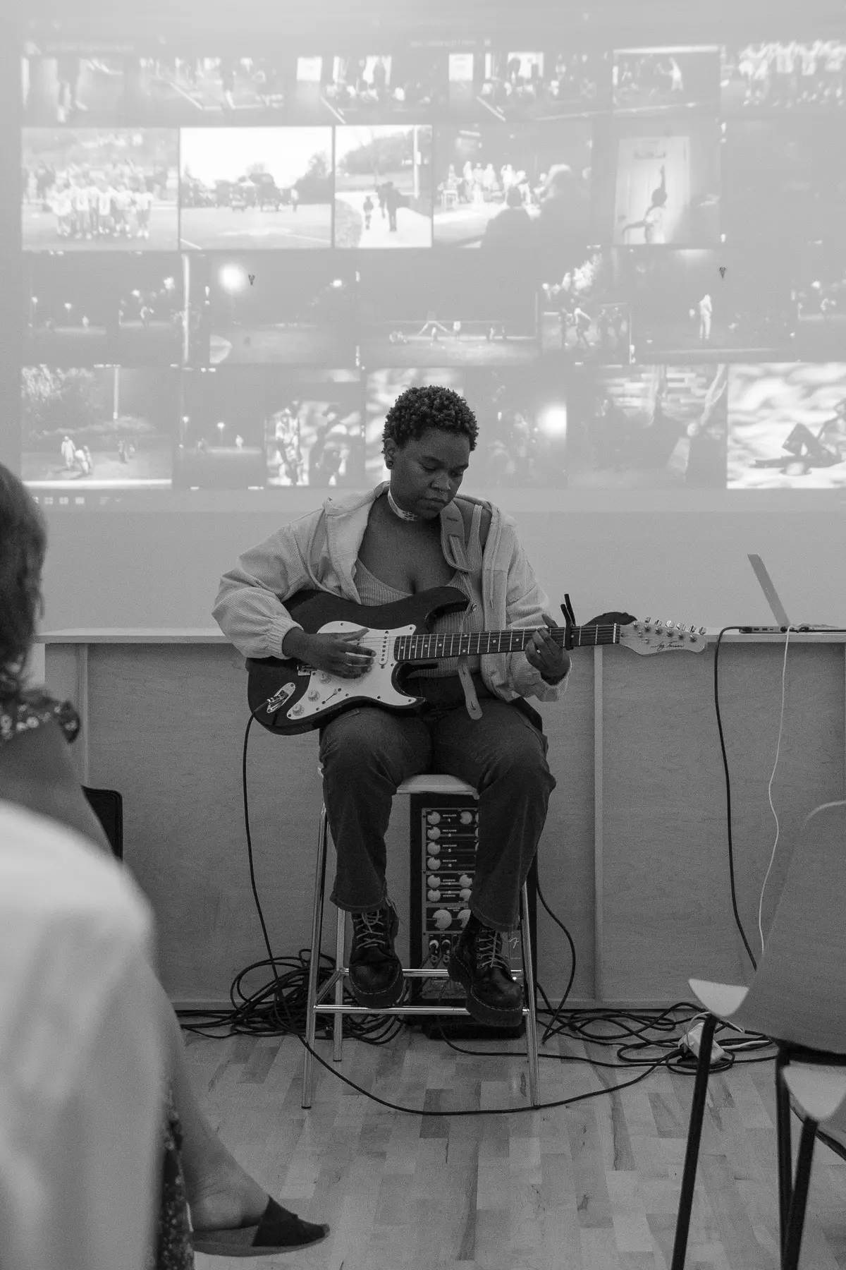 Musician Susa performs an intimate electric guitar set for a seated audience in a small, modern gallery space, with a projected black-and-white photo collage displayed behind them.