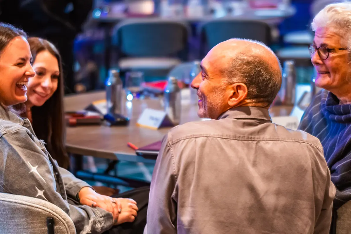 Four people sitting together around a table, smiling and engaged in lively conversation during a Salon, with notebooks, water bottles, and name cards visible on the table in a warmly lit setting.