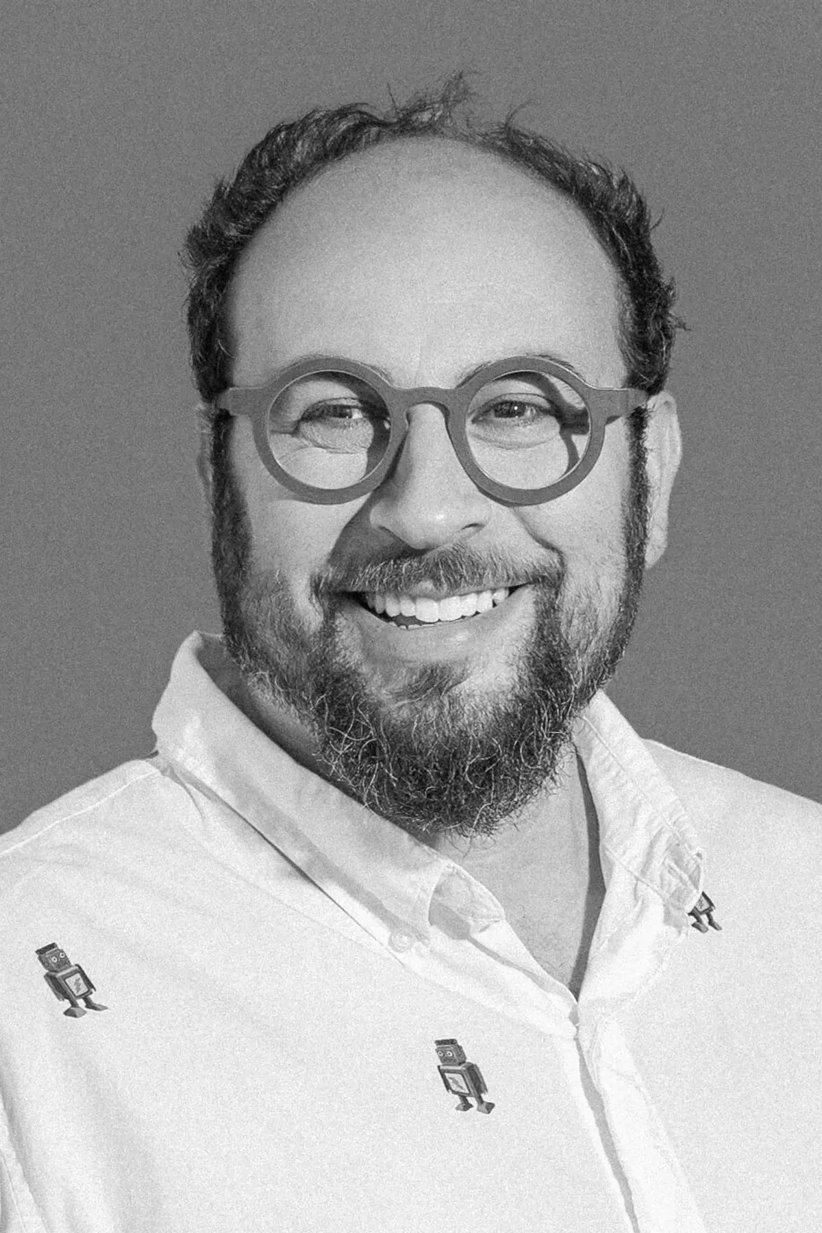Seth Goldenberg standing against a plain background, smiling warmly, wearing round glasses and a light-colored button-down shirt with a playful pattern, captured in a black-and-white portrait.
