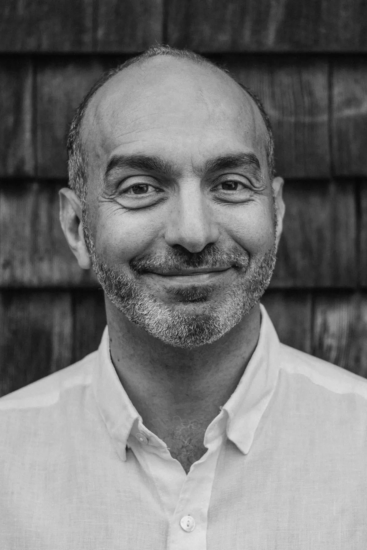Smiling portrait of Alex Jadad wearing a light-colored button-down shirt, standing in front of a textured wooden shingle wall.