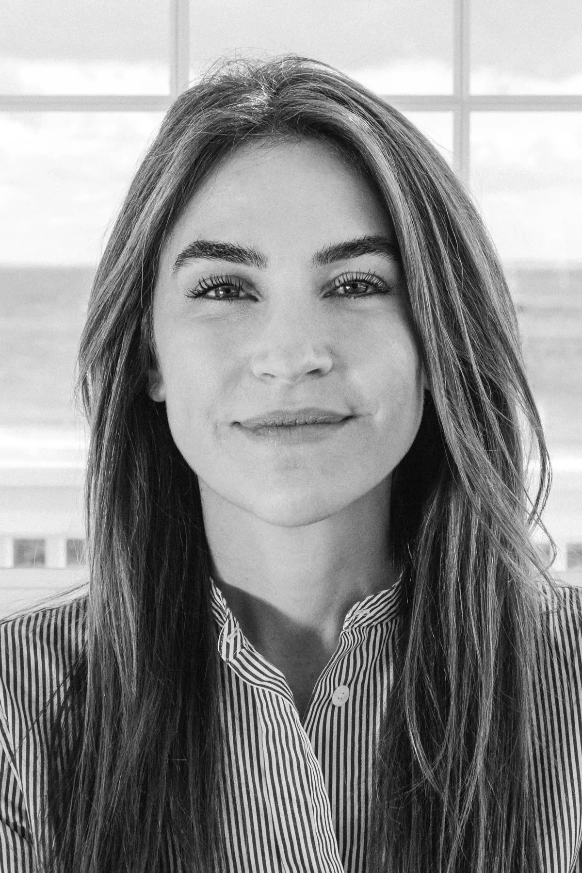 Morgwn Rimel seated on a couch in front of a large window with an ocean view, wearing a striped button-down shirt and smiling gently in natural light.